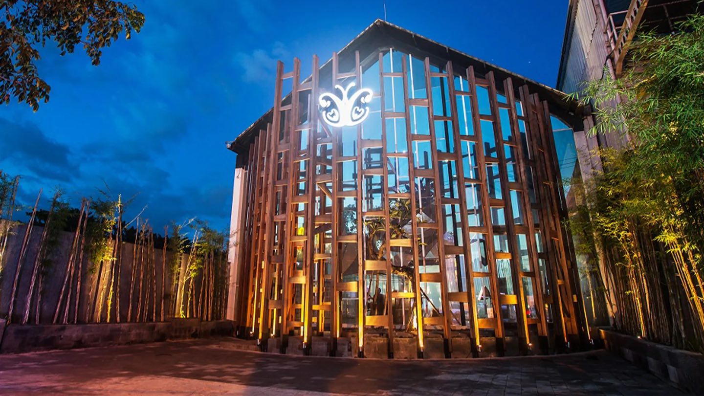 The Cocoon Legian building at dusk, its tall timber lattice lit gold beneath the butterfly logo