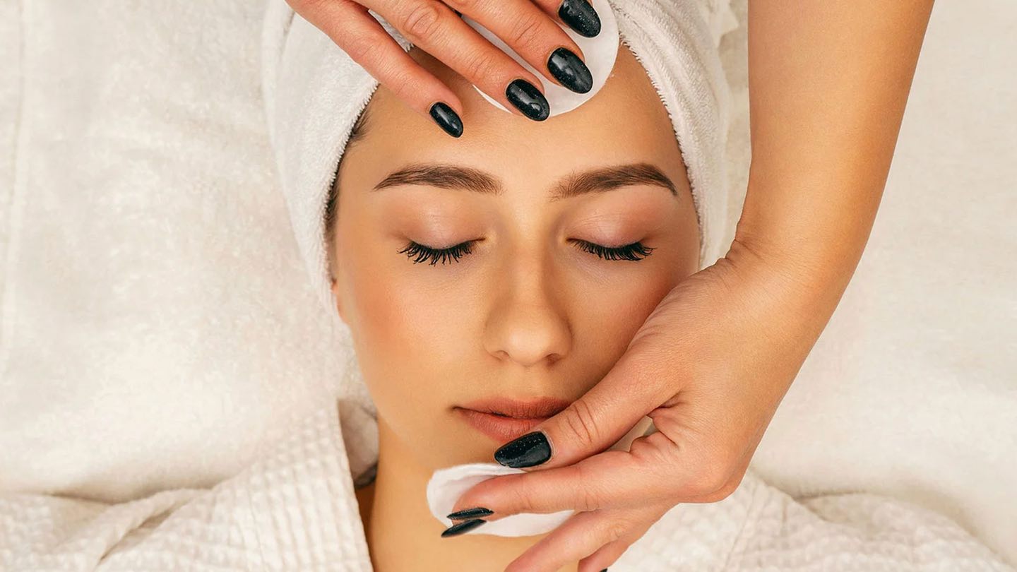 A therapist applying a peel to a woman’s face as she rests with her eyes closed, hair wrapped in a towel