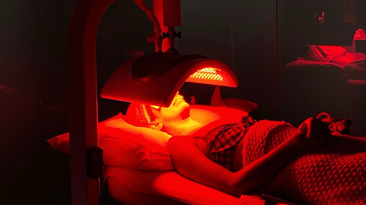 A guest resting under a red LED light panel in a dark Cocoon treatment room