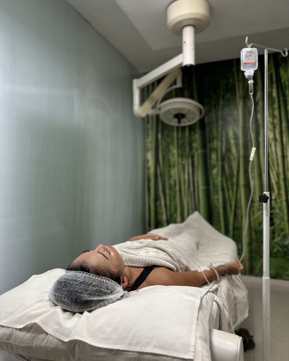 A guest resting under a blanket while a drip runs, in front of the bamboo wall of the Legian drip lounge