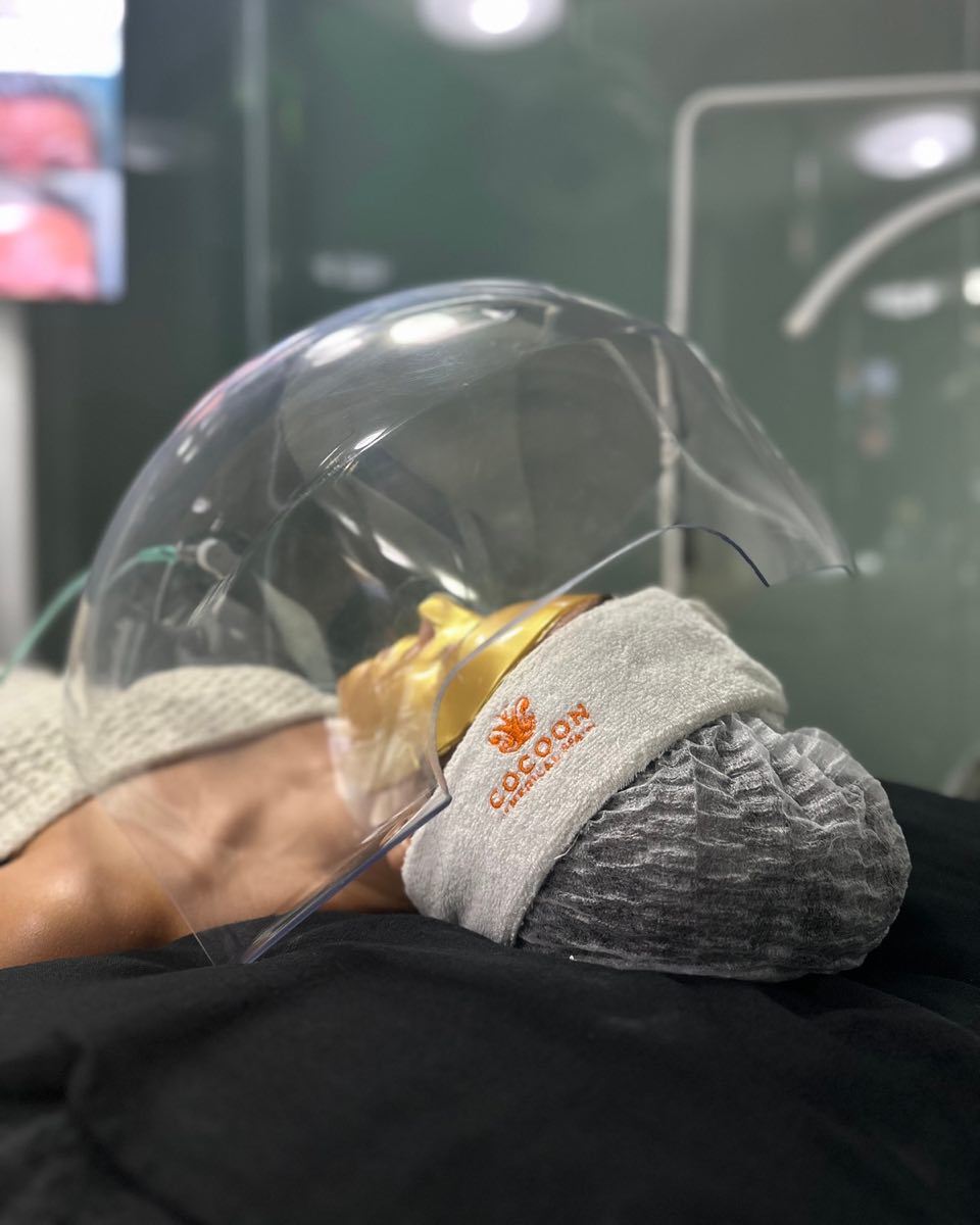 A guest resting under the oxygen dome in a gold mask, a Cocoon headband over her cap