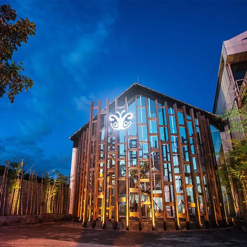 The Cocoon Legian building at dusk: a tall timber lattice lit gold, the butterfly logo glowing at the top