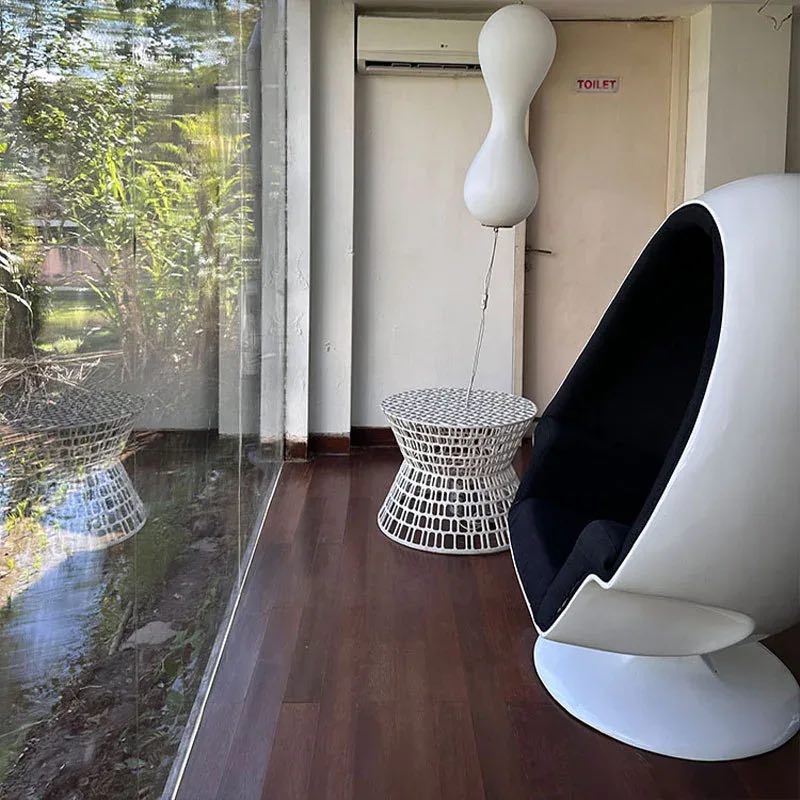 A black egg chair and a woven side table beside a glass wall onto tropical plants