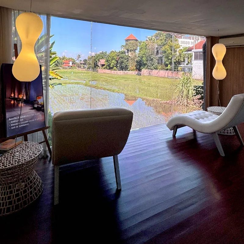 A lounge chair by a glass wall looking out over a green rice field, lit by two amber lamps