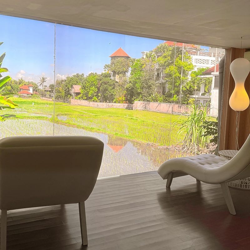 The Ubud lounge: a white armchair and chaise before a glass wall onto a green rice field
