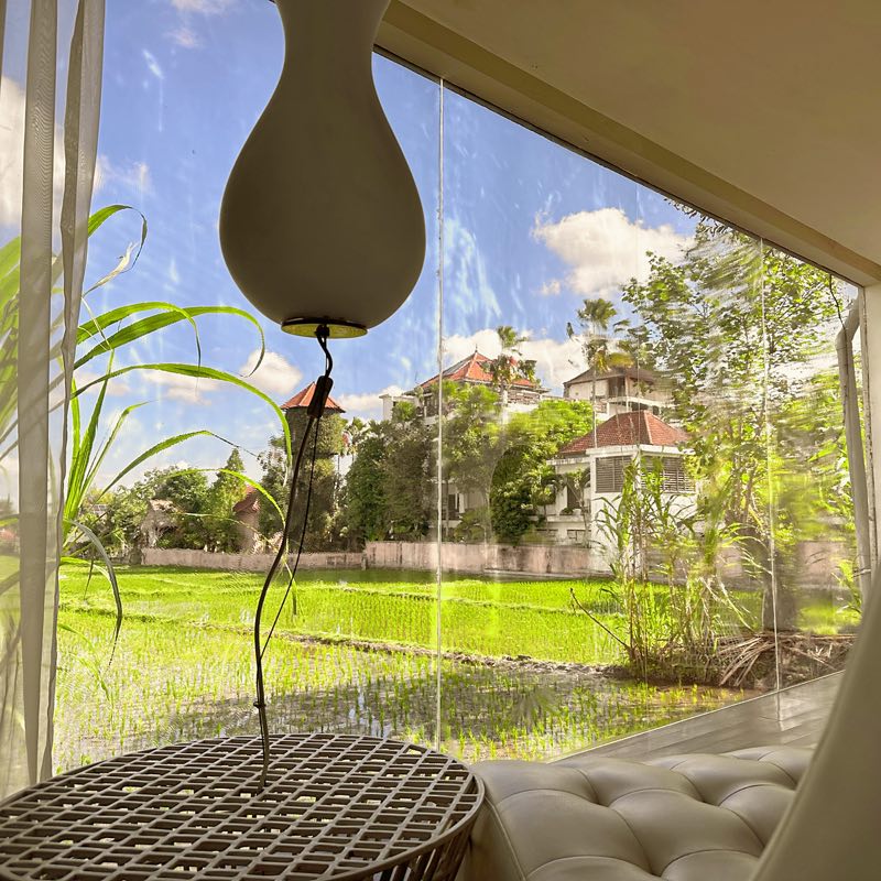 Looking out from the Ubud lounge: a white pendant lamp and a tufted chaise before a glass wall onto a green rice field
