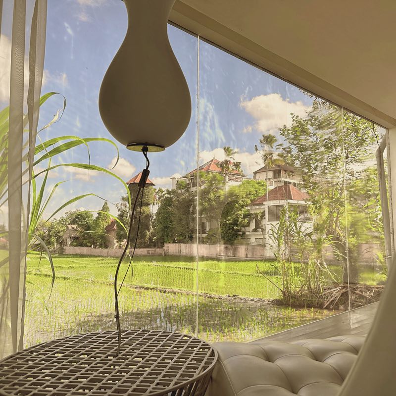 Looking out from the Ubud lounge: a white pendant lamp and a tufted chaise before a glass wall onto a green rice field