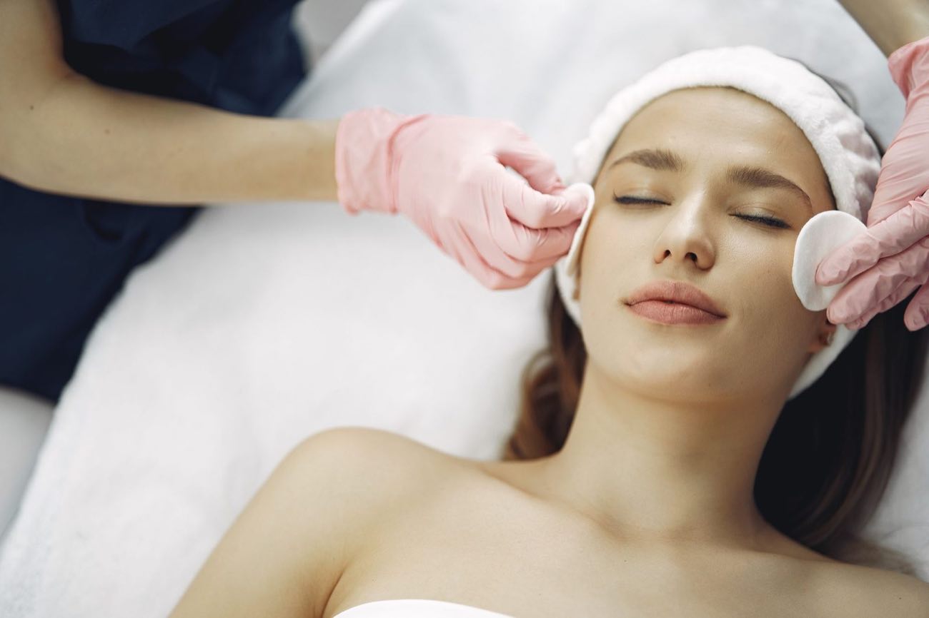 A woman in a white towel turban rests with her eyes closed while a clinician’s gloved hands touch lightly at her temples.