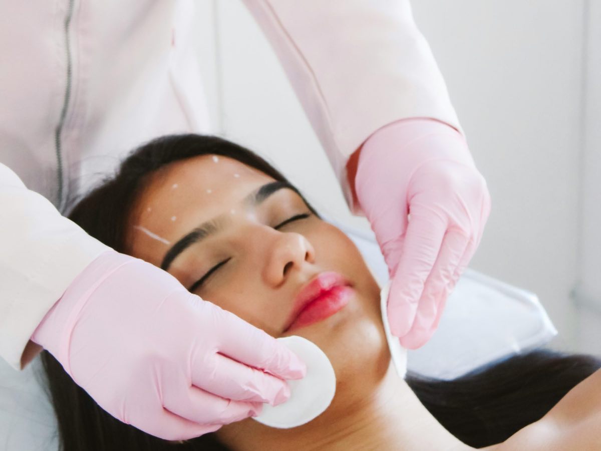 A woman rests with her eyes closed while gloved hands prepare her skin with cotton pads; small white points are marked on her forehead.