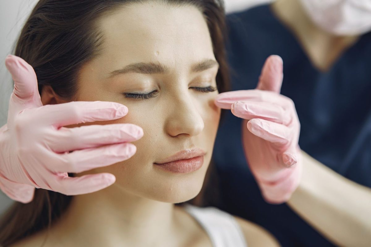 A woman with her eyes closed as a clinician’s pink-gloved fingertips rest lightly on both cheeks.