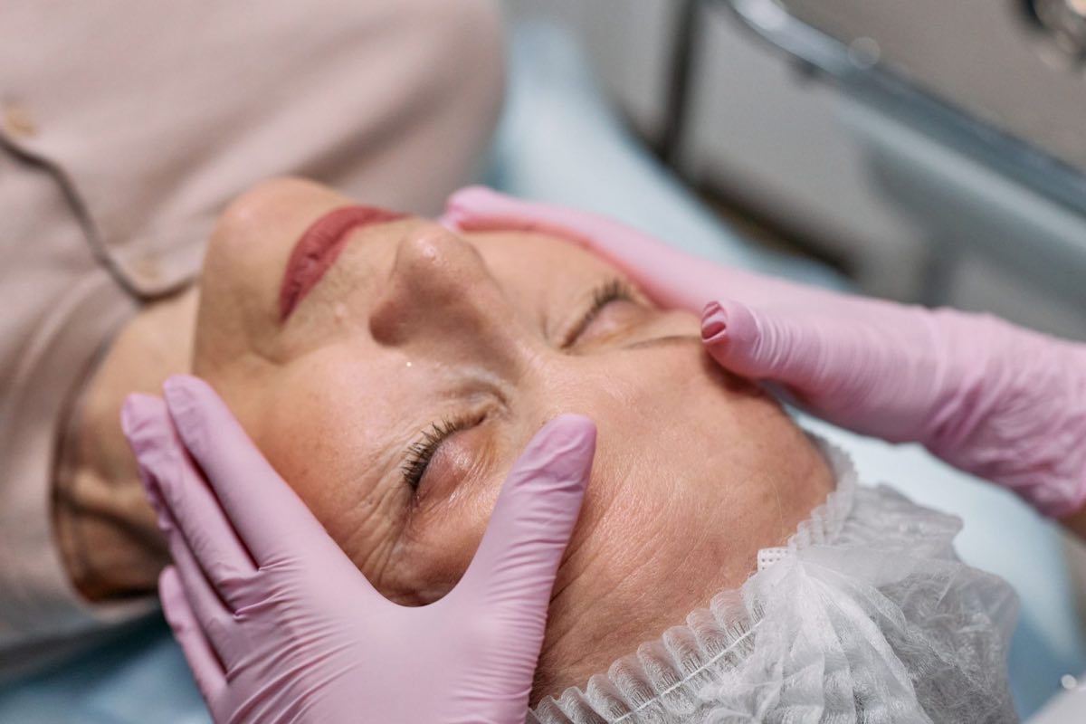 A woman in her sixties rests with her eyes closed as a clinician’s pink-gloved fingertips rest beside her eye and at her temple.